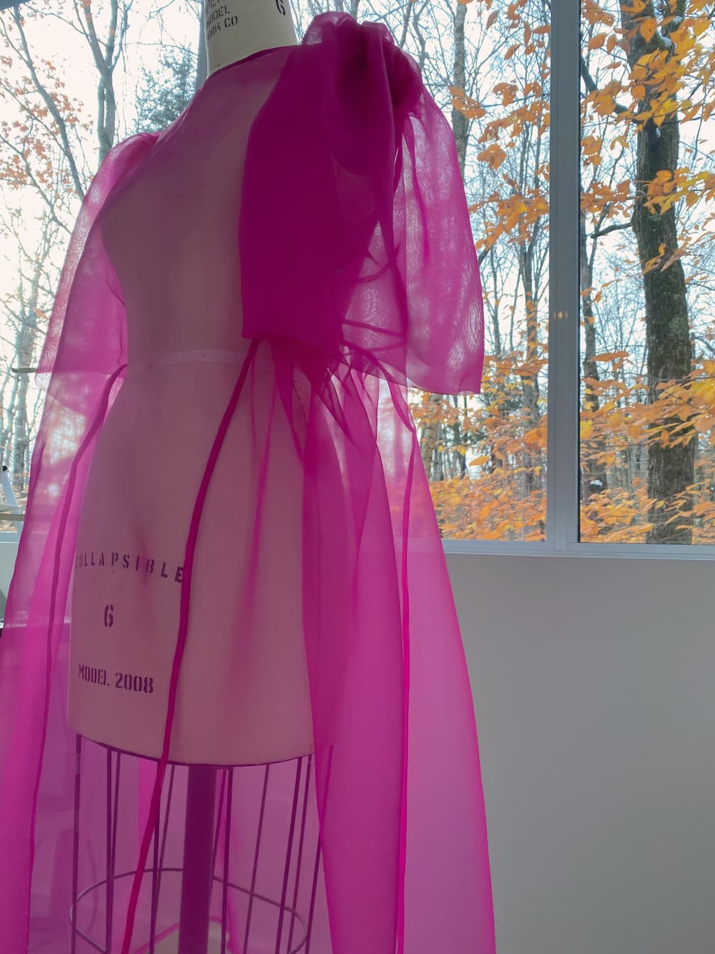 The Esperanza Midi Dress, crafted from vibrant pink organza silk by Noémiah, is elegantly displayed on a mannequin in front of a large window. Outside, the trees adorned with orange autumn leaves add a warm and inviting atmosphere.