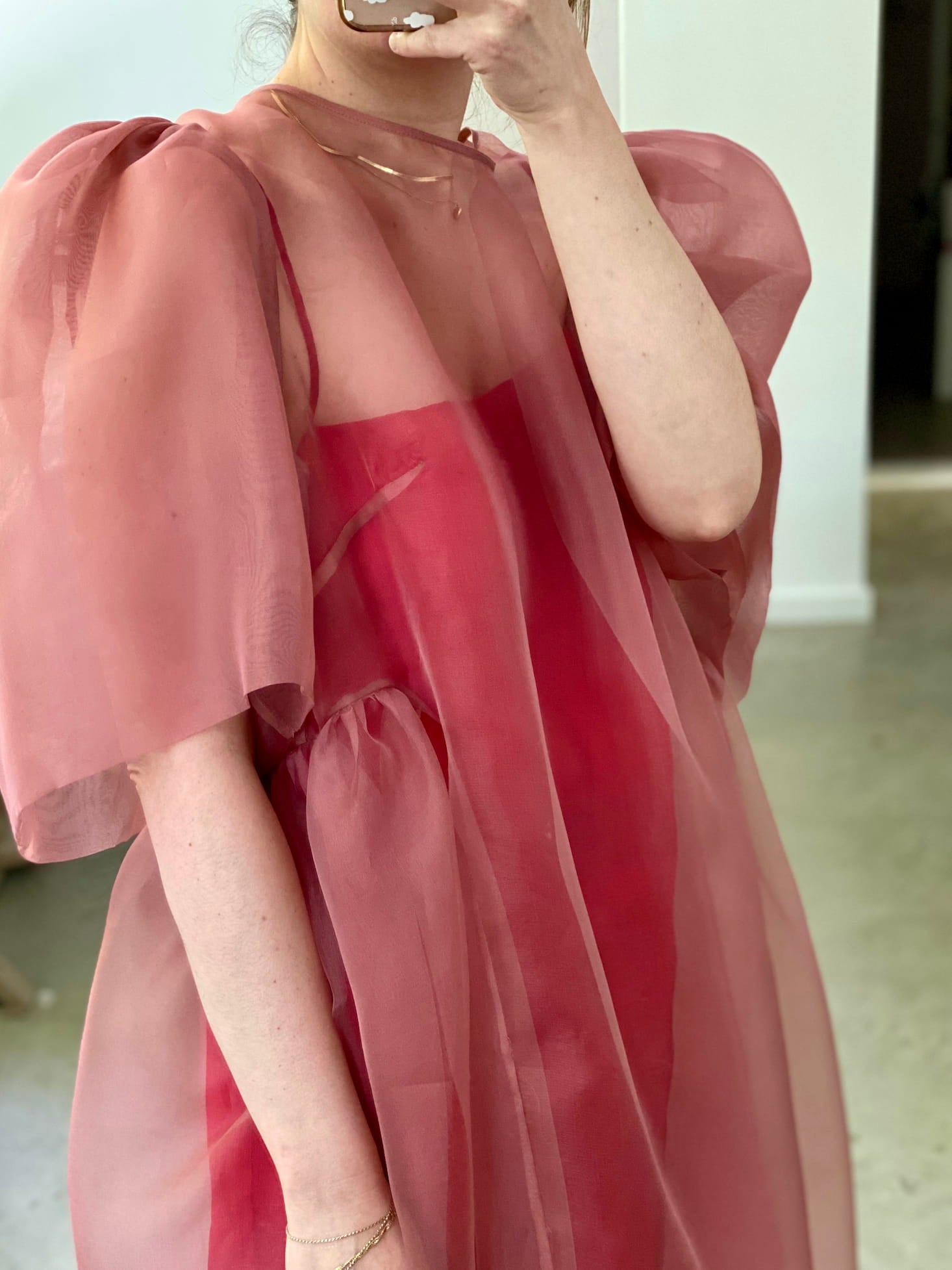 A person is taking a mirror selfie wearing the Esperanza Midi Dress — Organza Silk by Noémiah, featuring sheer, pink silk organza with puffed sleeves and a red underlayer. The setting is indoors with a neutral-toned background.