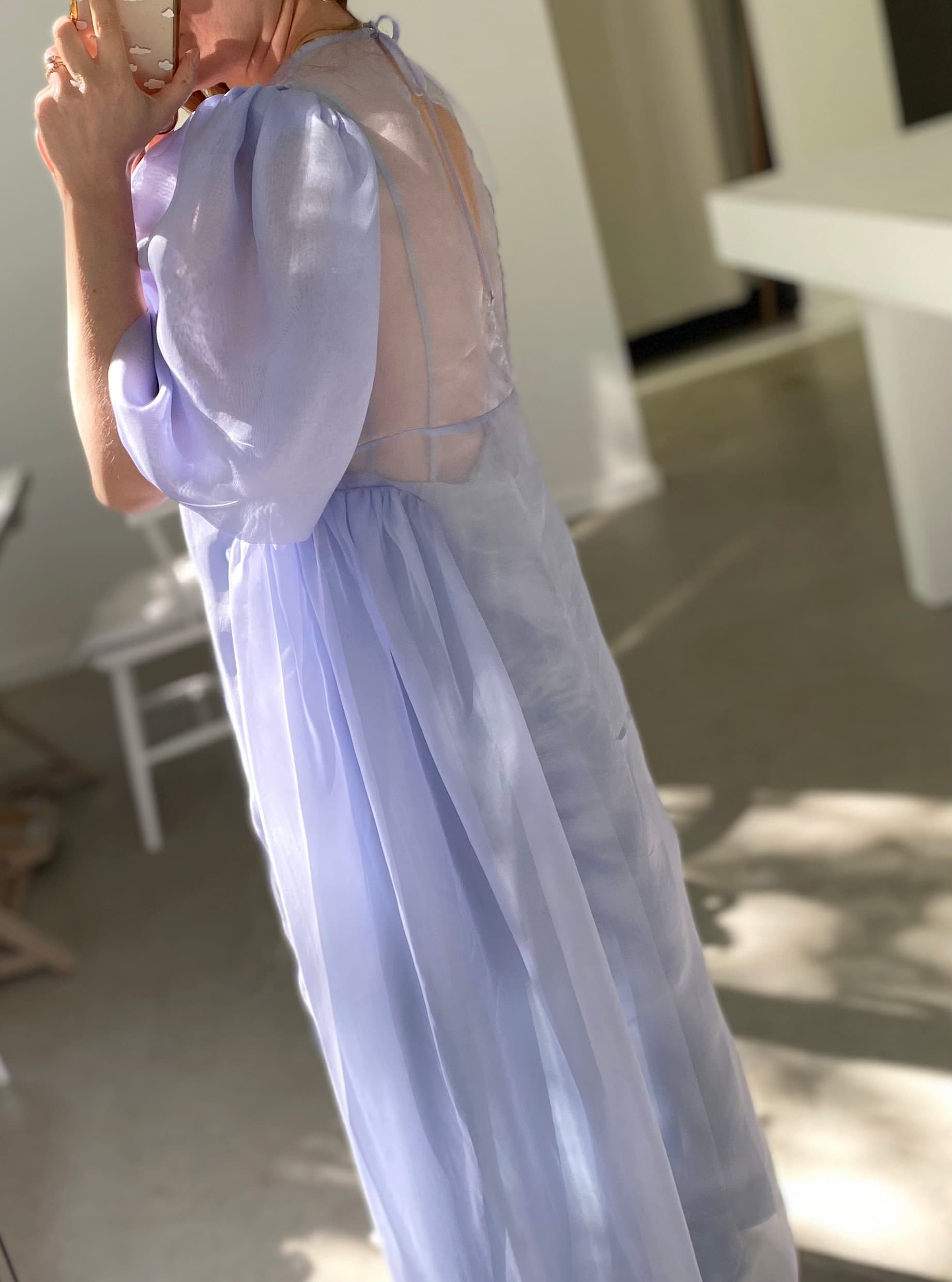 A person in the Esperanza Midi Dress — Organza Silk by Noémiah, featuring sheer fabric and puffed sleeves, holds a cup near their face. They stand in a sunlit indoor space with modern furnishings, including a white chair and table. Shadows and light create a soft ambiance.