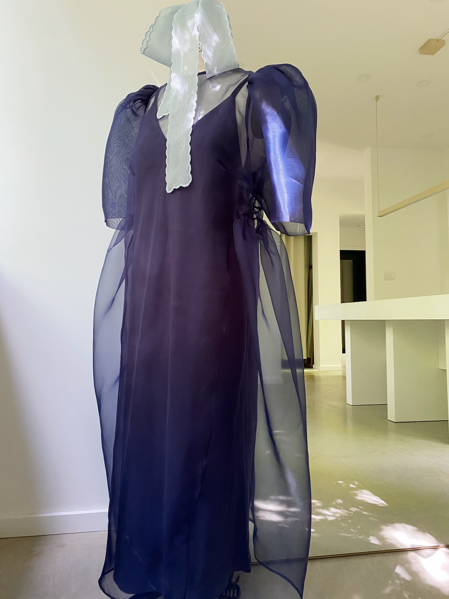 A mannequin showcases the Esperanza Midi Dress — Organza Silk by Noémiah, a semi-transparent dress in dark blue organza featuring puffed sleeves. The striking ensemble is complemented by a white textured ribbon draped over the face, all set against the minimalist room's light-colored walls and flooring for an elegant contrast.