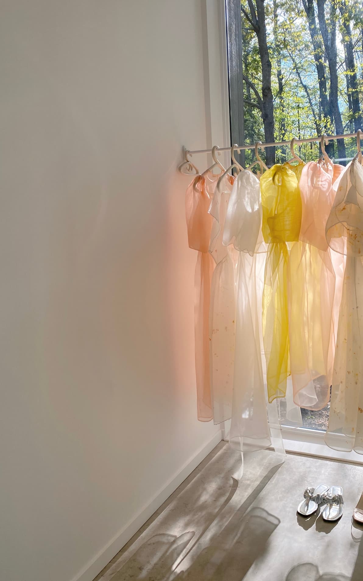 A sunlit room displays Noémiah's Esperanza Midi Dresses in pastel-colored organza silk, hanging neatly by a large window. These dresses, with their delicate puffed sleeves, are available in shades of pale pink and yellow. Sunlight creates shadows on the white wall, while a pair of sandals rests on the floor nearby.