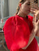 A person taking a mirror selfie is dressed in the Esperanza Midi Dress made of organza silk by Noémiah. The dress features bright red sheer material with elegant puffed sleeves. They hold a phone with a cloud-patterned case, their braided hair adorned with hoop earrings and several rings, while sunlight filters through the window behind them.