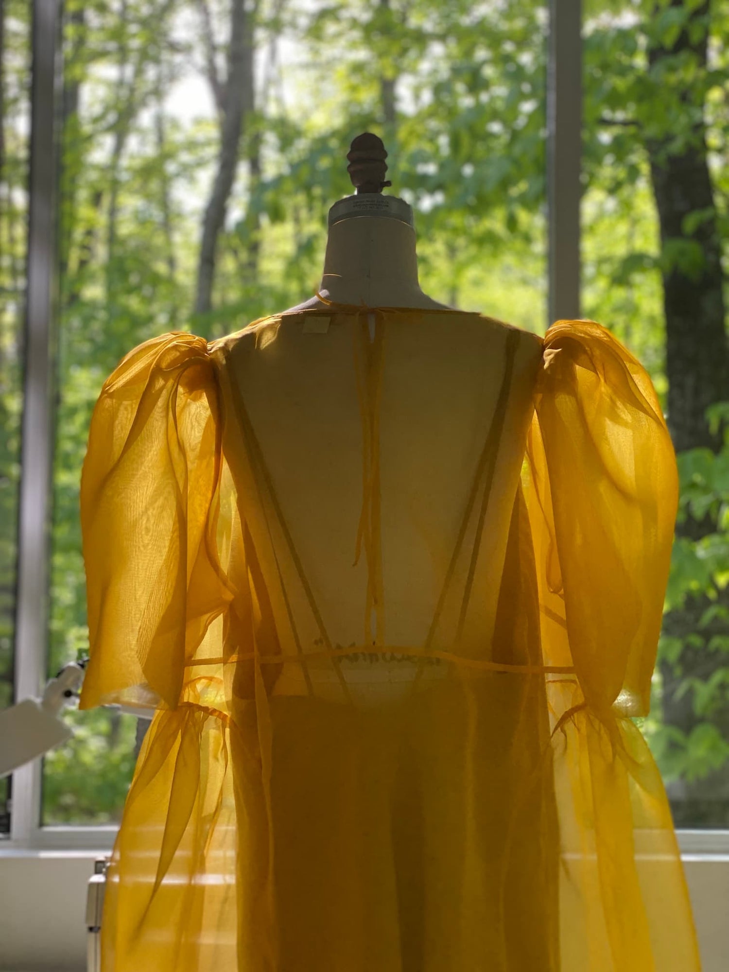 A mannequin showcases the Noémiah Esperanza Midi Dress—an organza silk design in sheer yellow with puffed sleeves and elegant cutout detail—in front of sunlit windows overlooking lush green trees.