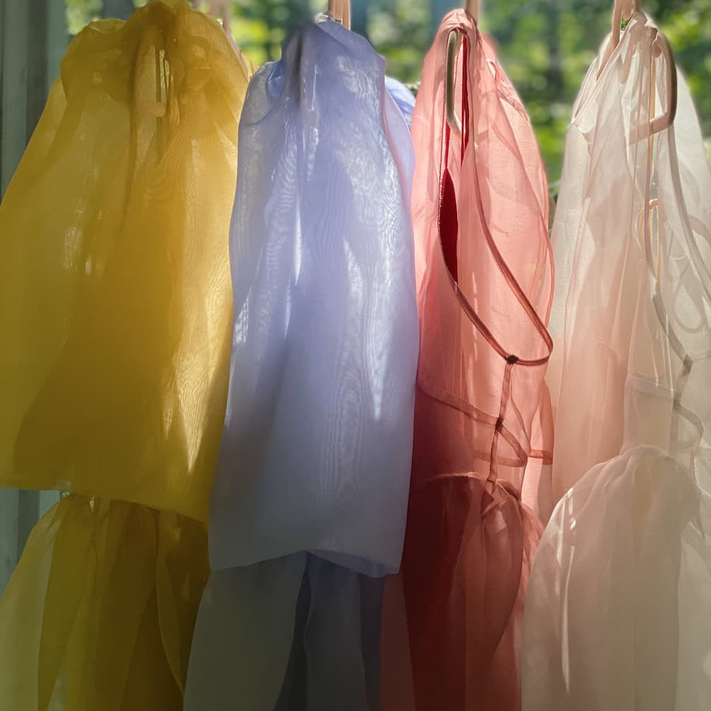Four sheer fabrics in yellow, lavender, pink, and peach hang side by side like the layers of the Esperanza Midi Dress — Organza Silk by Noémiah, backlit by sunlight streaming through a window with green trees outside.