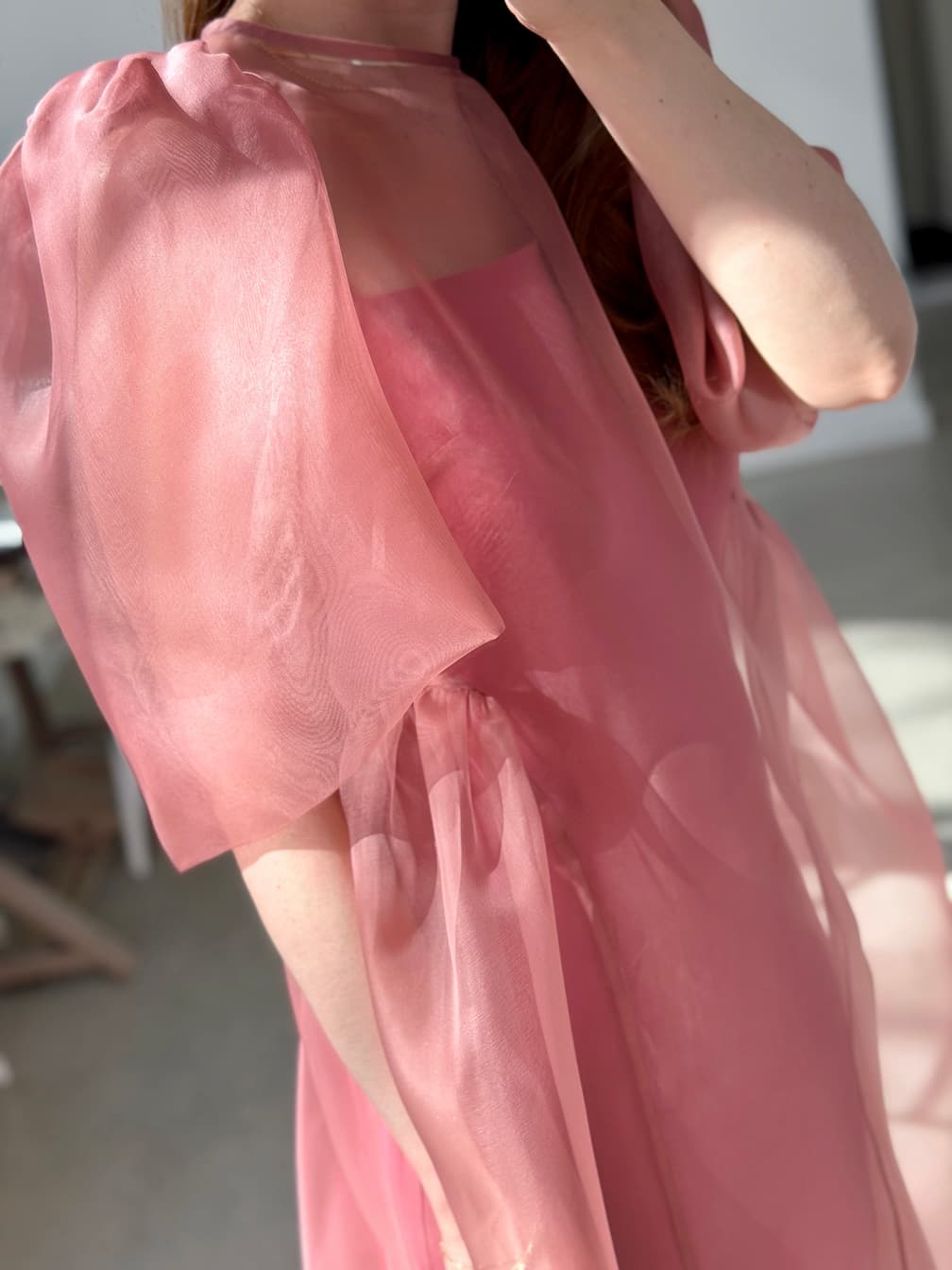 A person wears the Esperanza Midi Dress — Organza Silk by Noémiah, a sheer, flowing pink design with puffed sleeves. Only their upper body is visible in soft natural indoor light; their face is not shown.