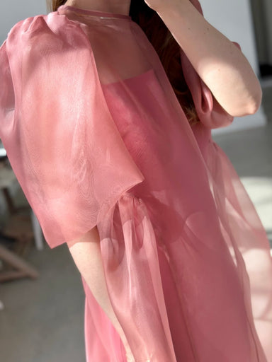 A person wears the Esperanza Midi Dress — Organza Silk by Noémiah, a sheer, flowing pink design with puffed sleeves. Only their upper body is visible in soft natural indoor light; their face is not shown.