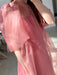 A person wears the Esperanza Midi Dress — Organza Silk by Noémiah, a sheer, flowing pink design with puffed sleeves. Only their upper body is visible in soft natural indoor light; their face is not shown.