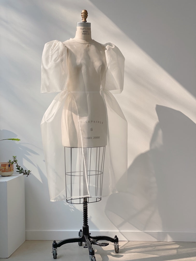 In a sunlit room, the Esperanza Midi Dress by Noémiah stands elegantly on a dress form, its sheer organza silk fabric with puffed sleeves catching the light. Shadows play across the white wall behind it, while a small plant sits gracefully on a nearby stand.