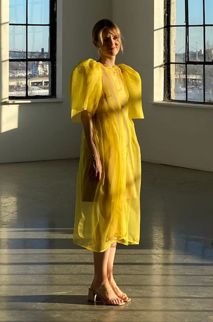 A person stands in a sunlit room wearing the elegant Esperanza Midi Dress by Noémiah, crafted from organza silk and featuring charming puffed sleeves. They are barefoot, with the sunlight casting shadows on the floor, while large windows frame an urban skyline outside.