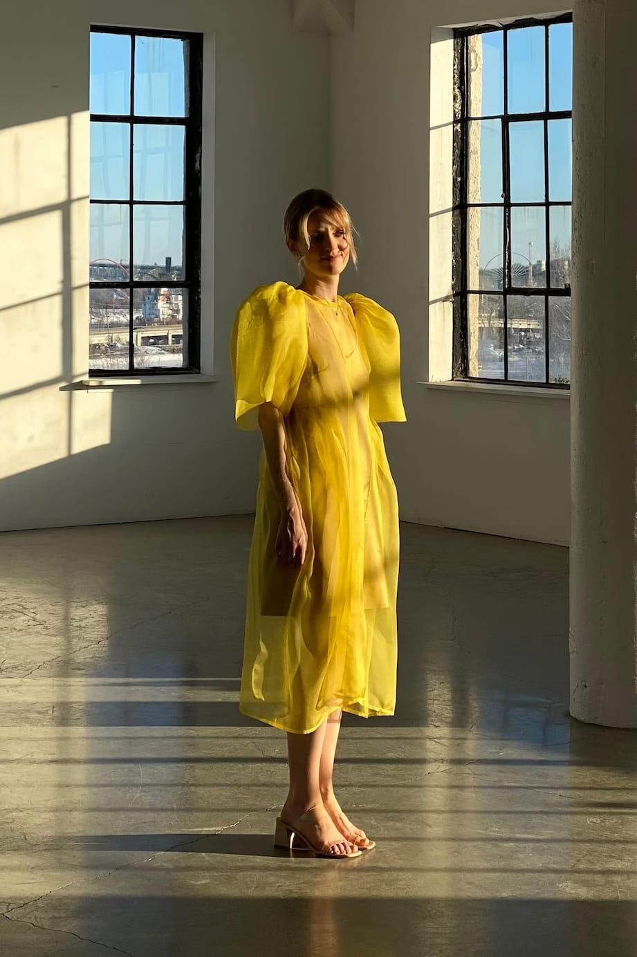 A person stands in a sunlit room wearing the elegant Esperanza Midi Dress by Noémiah, crafted from organza silk and featuring charming puffed sleeves. They are barefoot, with the sunlight casting shadows on the floor, while large windows frame an urban skyline outside.