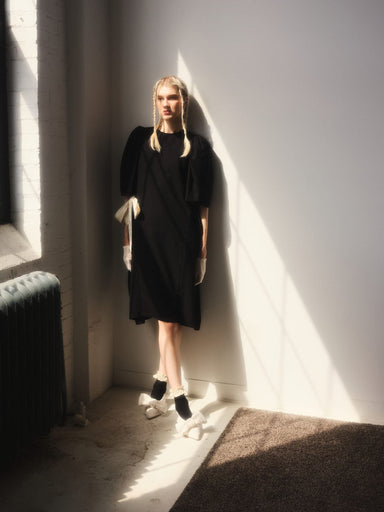 In a sunlit room, a woman leans against a white wall showcasing the exquisite Esperanza Midi Dress in raw silk by Noémiah. She pairs it elegantly with white gloves and distinctive black shoes adorned with large white bows. Soft shadows from a window cast geometric patterns across the floor and wall.