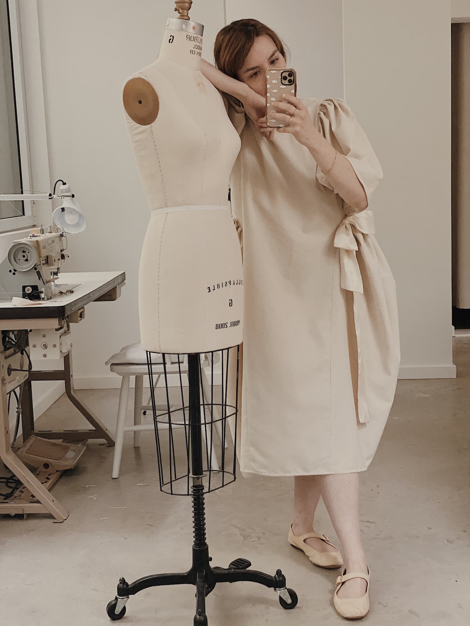 A person in a dress reminiscent of the Noémiah Esperanza Midi Dress — Raw Silk poses while leaning against a dress form in a sewing studio. They hold a smartphone to capture a mirror selfie, with the background showcasing a sewing machine and table, completing the creative scene.