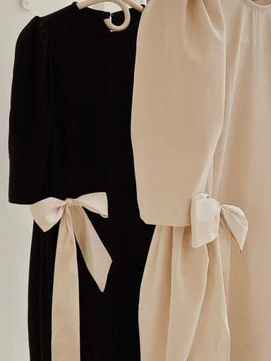 Hanging on the rack are two Esperanza Midi Dresses by Noémiah: one in classic black raw silk and the other in beige. Both dresses are adorned with puffed sleeves and matching fabric bows at the waist, offering a stylish and elegant appearance.