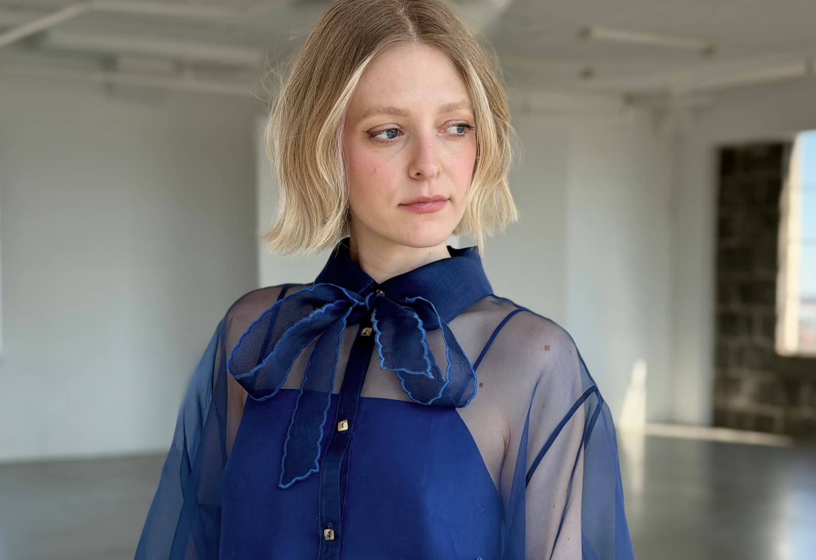 A plus size woman with short blonde hair models the Flavie Shirt Dress — Classic by Noémiah, an oversized blue silk organza blouse with a large bow, standing thoughtfully in a bright, empty room.