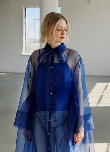 A woman with blond hair stands indoors wearing the Noémiah Flavie Shirt Dress — Classic in sheer blue organza over a blue top and jeans, looking down as sunlight streams through the window behind her.