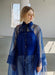 A woman with blond hair stands indoors wearing the Noémiah Flavie Shirt Dress — Classic in sheer blue organza over a blue top and jeans, looking down as sunlight streams through the window behind her.