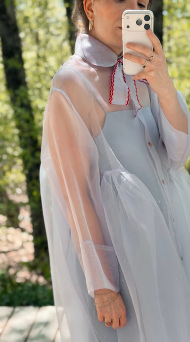 A person takes a mirror selfie outdoors in the Noémiah Flavie Shirt Dress — Classic, a sheer, oversized silk organza maxi with a high collar and red trim in light lavender. Blurred green trees are visible in the background.