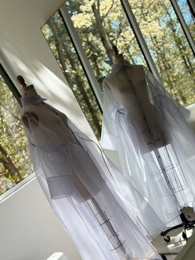 Two dress forms showcase Noémiah’s Flavie Shirt Dress — Classic in sheer white silk organza, displayed in a sunlit room with large windows revealing trees outside.