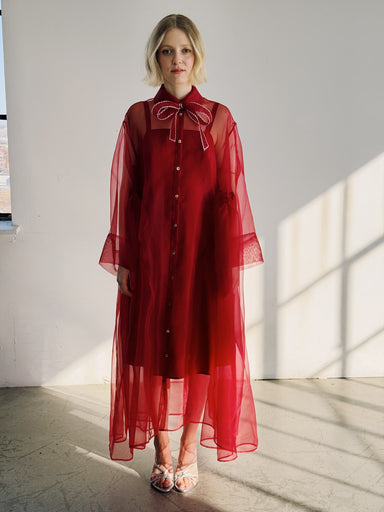 A woman stands indoors in the Flavie Shirt Dress — Classic by Noémiah, an oversized, sheer red maxi dress with bell sleeves and a large collar bow. She has short blonde hair and silver sandals as sunlight streams in, casting floor shadows.