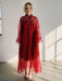 A woman stands indoors in the Flavie Shirt Dress — Classic by Noémiah, an oversized, sheer red maxi dress with bell sleeves and a large collar bow. She has short blonde hair and silver sandals as sunlight streams in, casting floor shadows.