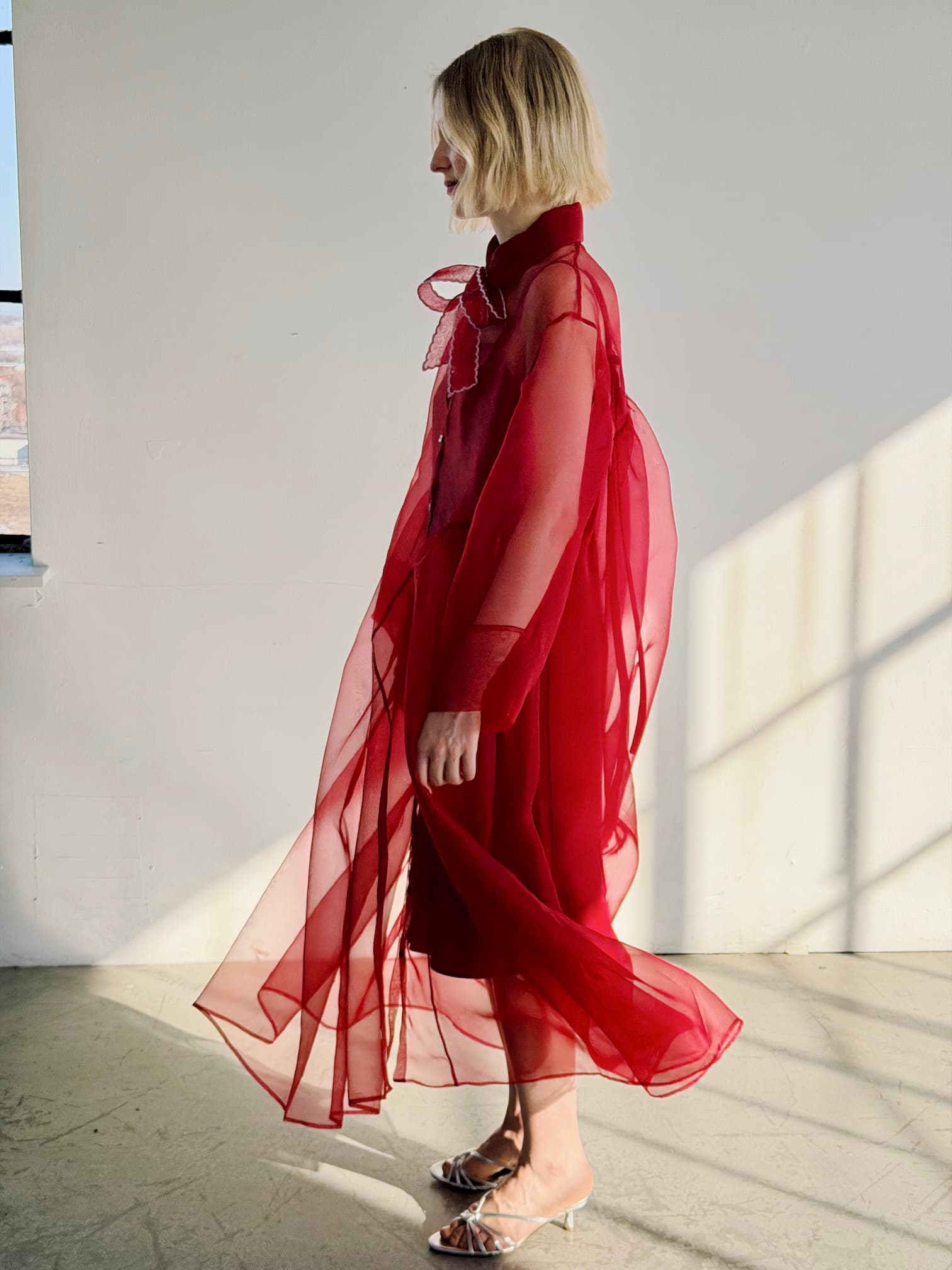 A woman with short blonde hair stands sideways in a sunlit room, wearing Noémiah's Silk Ribbon — Thin, styled as a bow at her collar. Soft light casts gentle shadows on the white wall behind her.