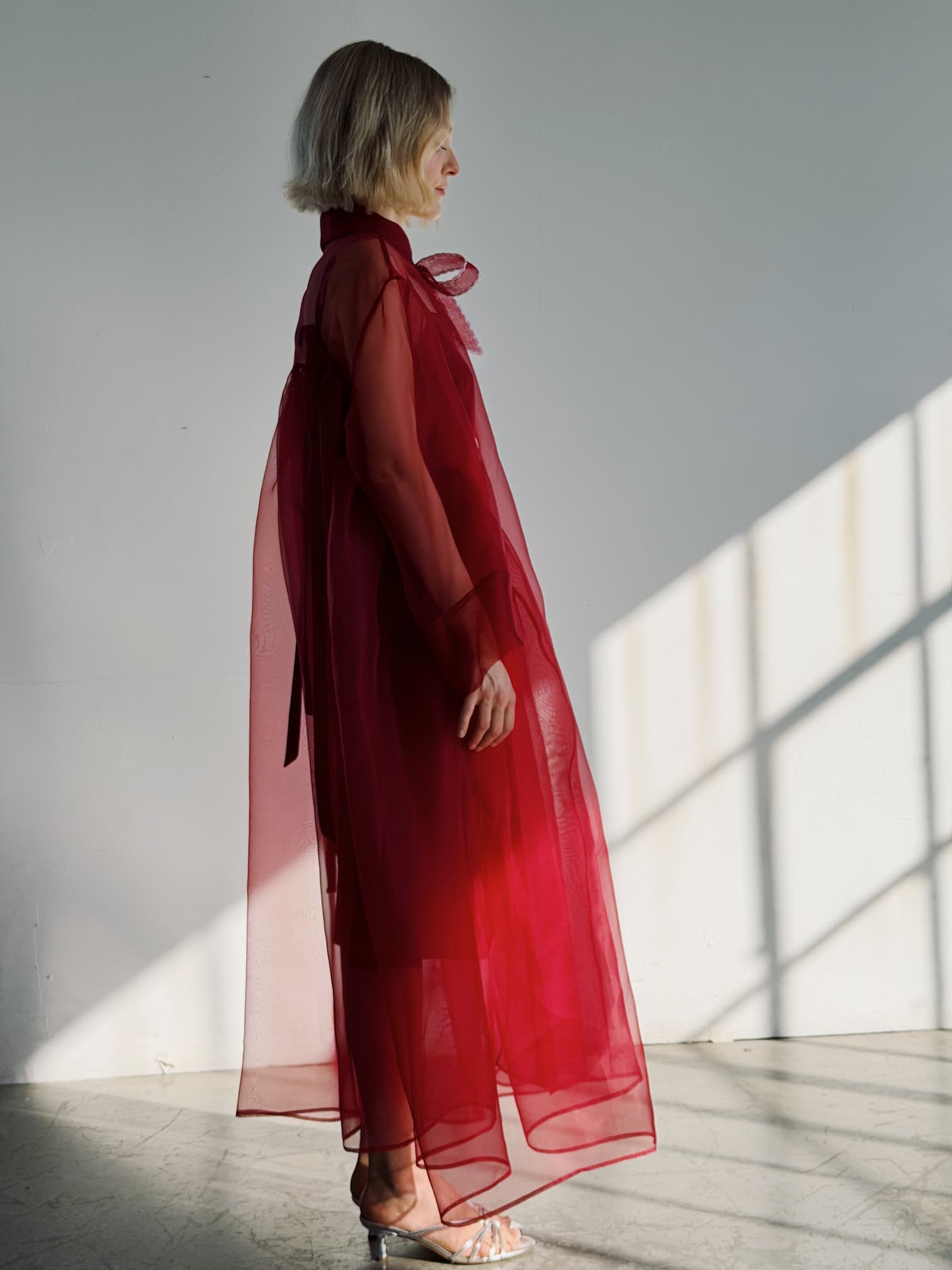 A person with short blonde hair stands in profile wearing the Noémiah Flavie Shirt Dress — Classic, a sheer, flowing red maxi dress with long sleeves and a neck bow, plus silver sandals. Sunlight streams through a window, casting shadows on the white wall and floor.