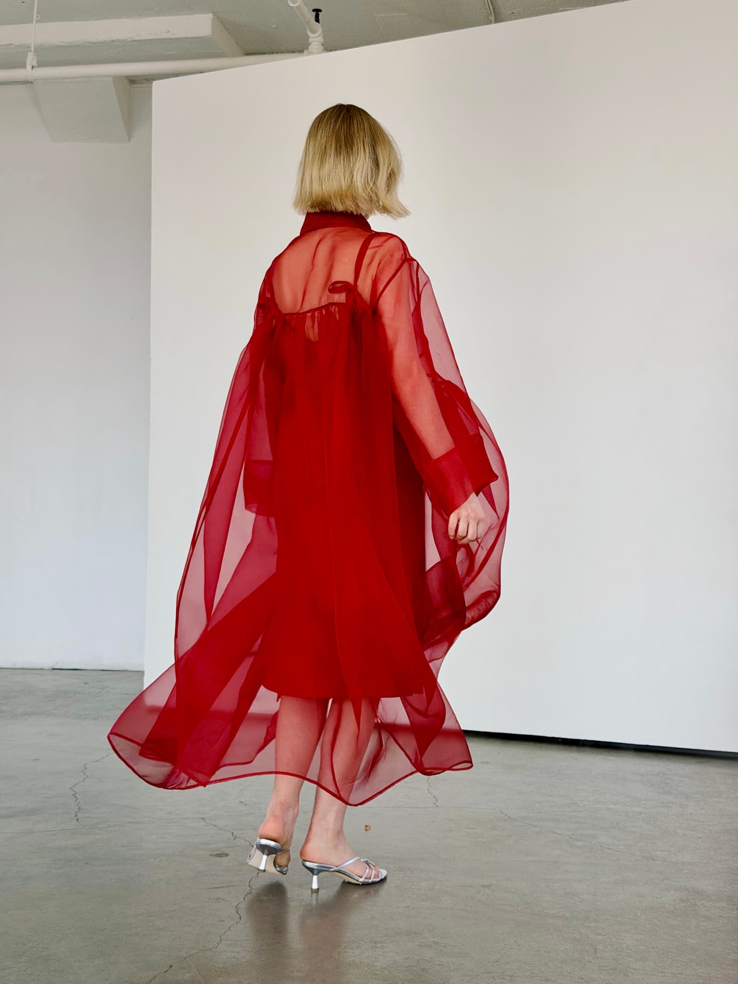 A person with short blonde hair, wearing the Noémiah Flavie Shirt Dress — Classic in red and silver heels, stands on a concrete floor in a minimalist white-walled room, facing away from the camera.