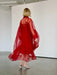 A person with short blonde hair, wearing the Noémiah Flavie Shirt Dress — Classic in red and silver heels, stands on a concrete floor in a minimalist white-walled room, facing away from the camera.