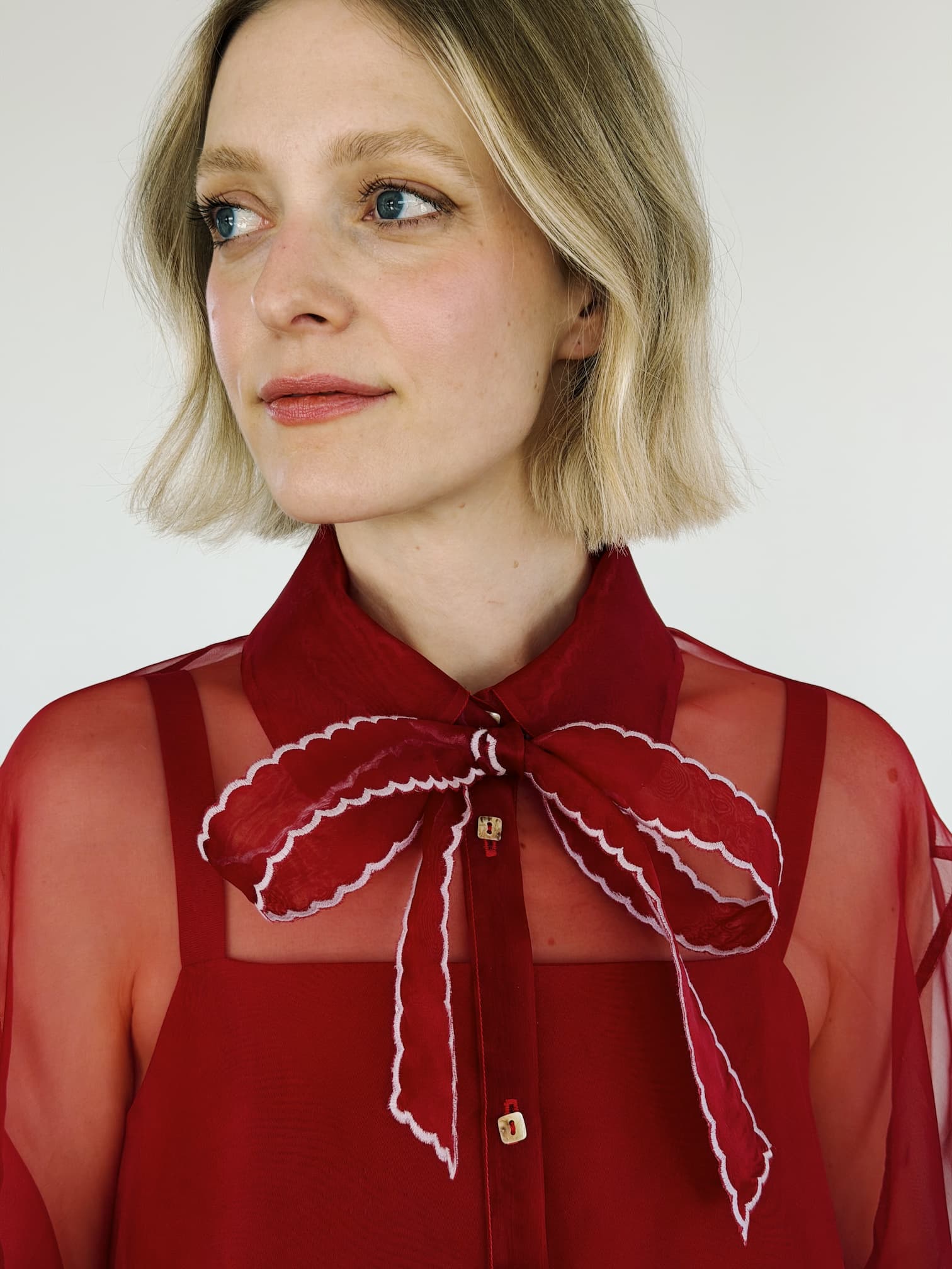A woman with short blonde hair wears the Flavie Shirt Dress — Classic by Noémiah, featuring a sheer red blouse with a large red bow and white trim over an oversized organza maxi dress, looking slightly to the side against a light background.