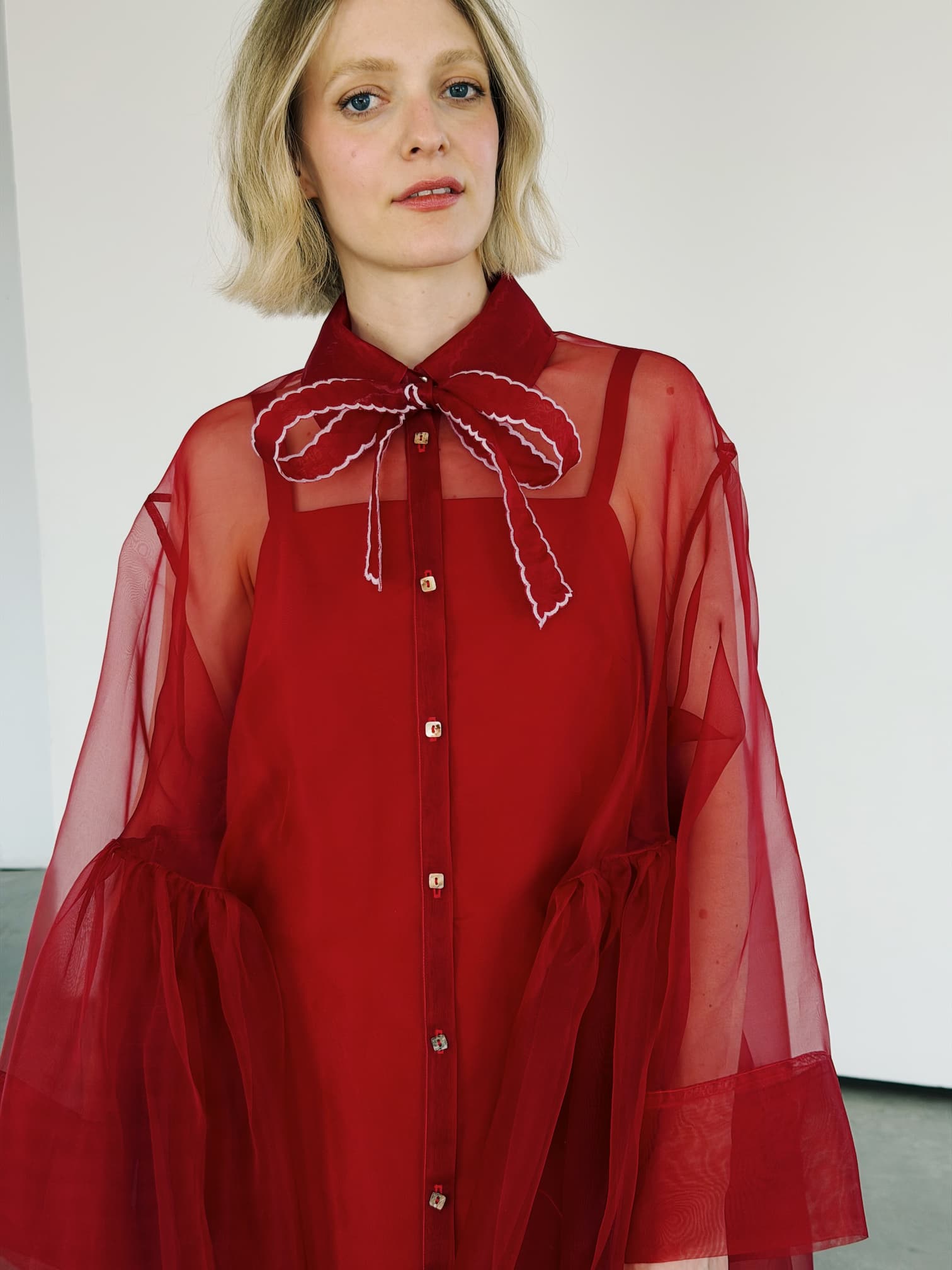 A woman with short blonde hair wears a Noémiah Silk Ribbon — Thin in red, styled as a bow at her collar, standing against a plain white background.