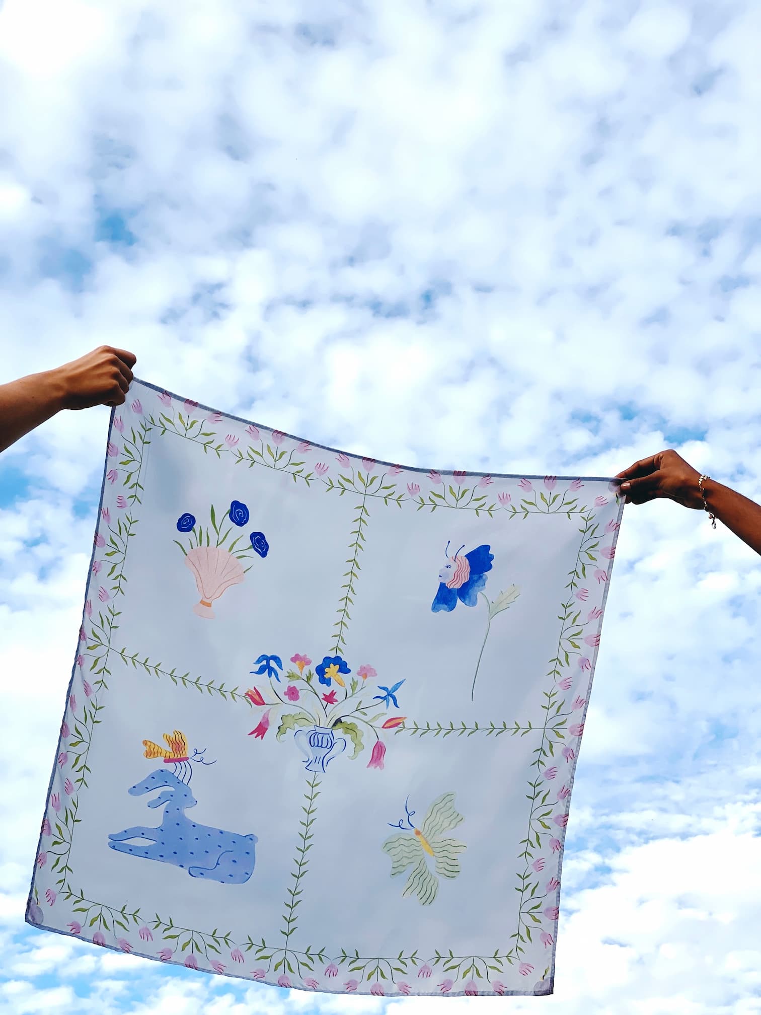 Two hands hold the Fleurs Coupées Scarf by Noémiah, showcasing its enchanting folk art style against a cloudy blue sky. Reminiscent of Estée Preda's work, this decorative silk scarf features animal and floral patterns within bordered sections, adding a whimsical touch to the scene.