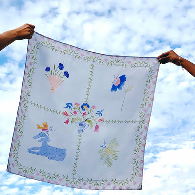 Two hands hold the Fleurs Coupées Scarf by Noémiah, showcasing its enchanting folk art style against a cloudy blue sky. Reminiscent of Estée Preda's work, this decorative silk scarf features animal and floral patterns within bordered sections, adding a whimsical touch to the scene.