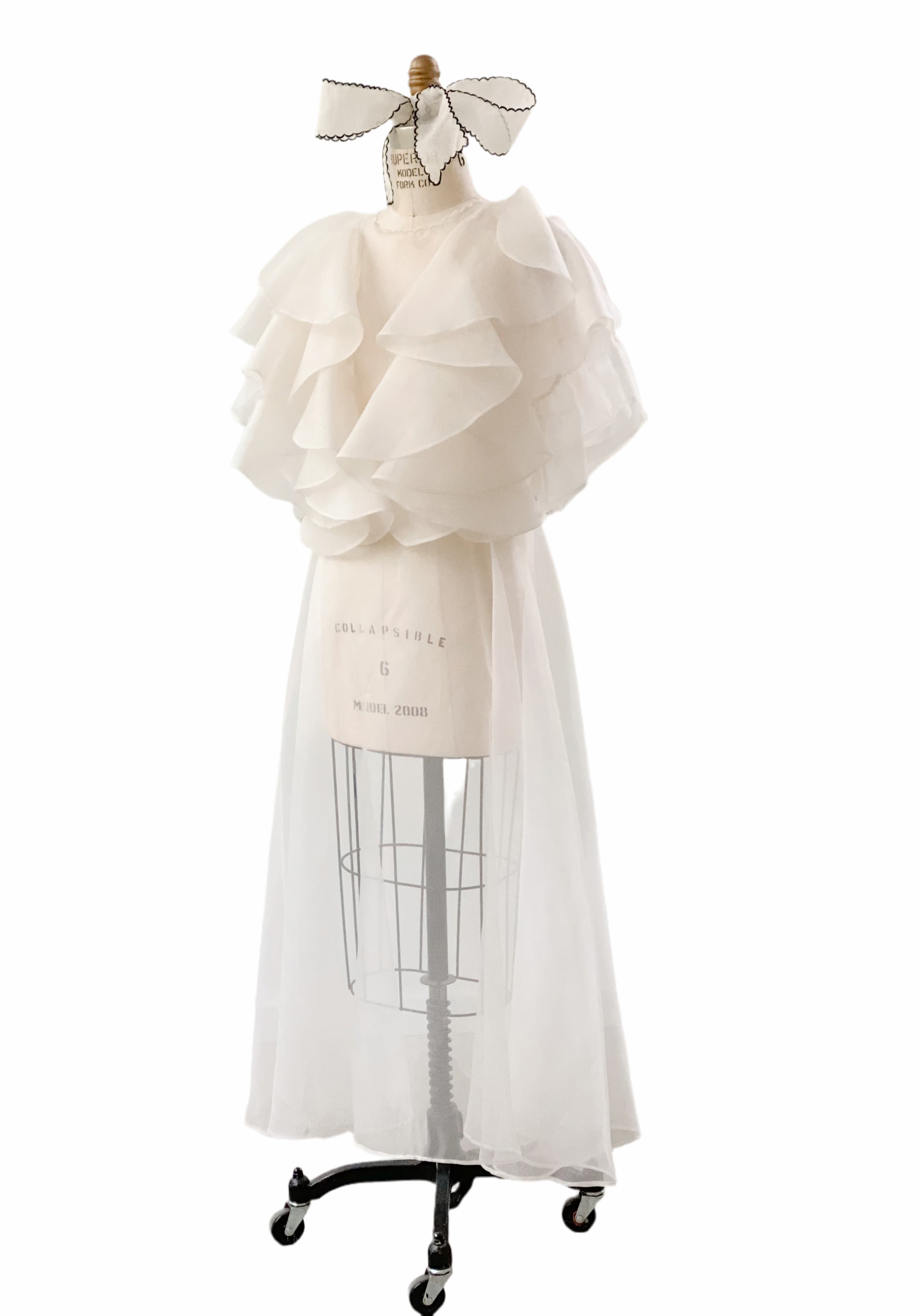 The Flora Dress by Noémiah is elegantly showcased on a dressmaker's mannequin, featuring a flowing, layered white design with ruffled details and a sheer silk organza overlay. This handmade-to-order piece is accentuated with an embroidered neckline and topped off with a decorative ribbon.