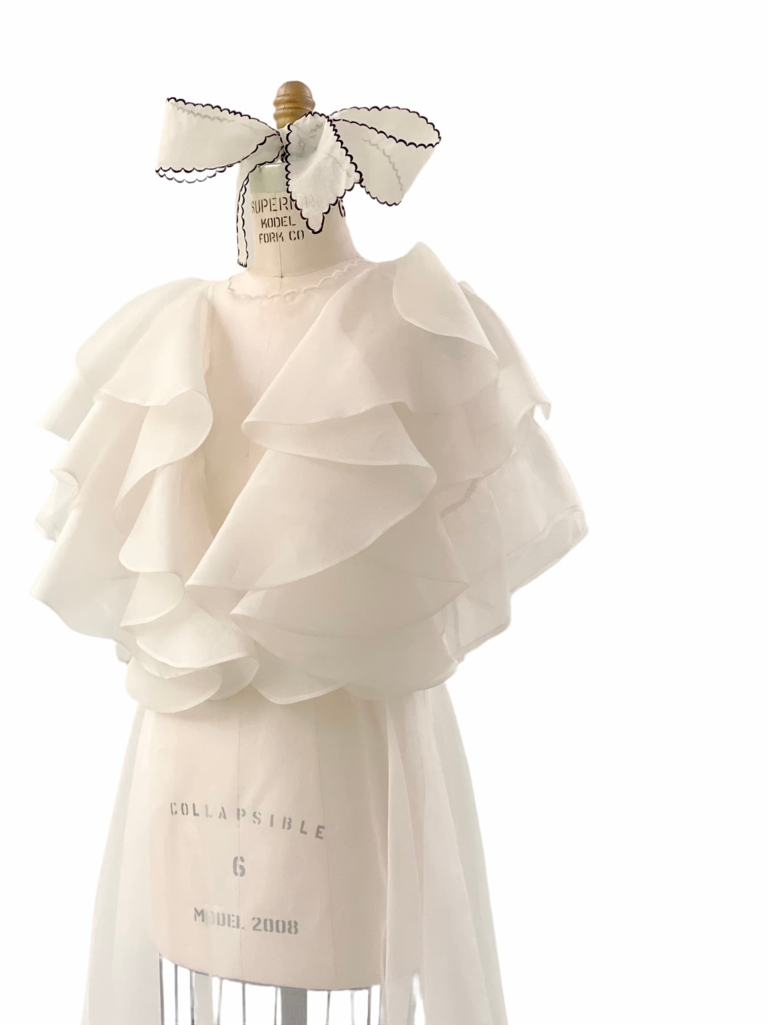 A dress form showcases the Flora Dress by Noémiah, a handcrafted white chiffon piece with tiered layers, a sheer finish, and an embroidered neckline. The display includes a fabric label featuring sewing machine stitching and is topped with a decorative mannequin head cover.