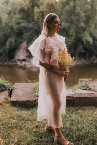 Wearing the Flora Dress by Noémiah, a woman in a sheer, light pink ensemble with an embroidered neckline holds a bouquet of yellow flowers. She stands on grass beside a calm river, surrounded by lush green trees, creating a serene and natural atmosphere.