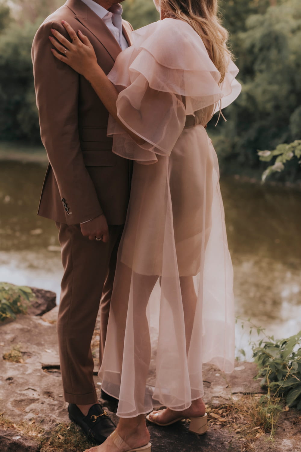 A couple stands by a serene pond, embraced by the lush greenery around them. The woman is elegantly styled in the Flora Dress from Noémiah, featuring sheer ruffles and an embroidered neckline, paired with heels. The man complements her aesthetic in a brown suit and black loafers. Their close embrace sets a romantic tone.