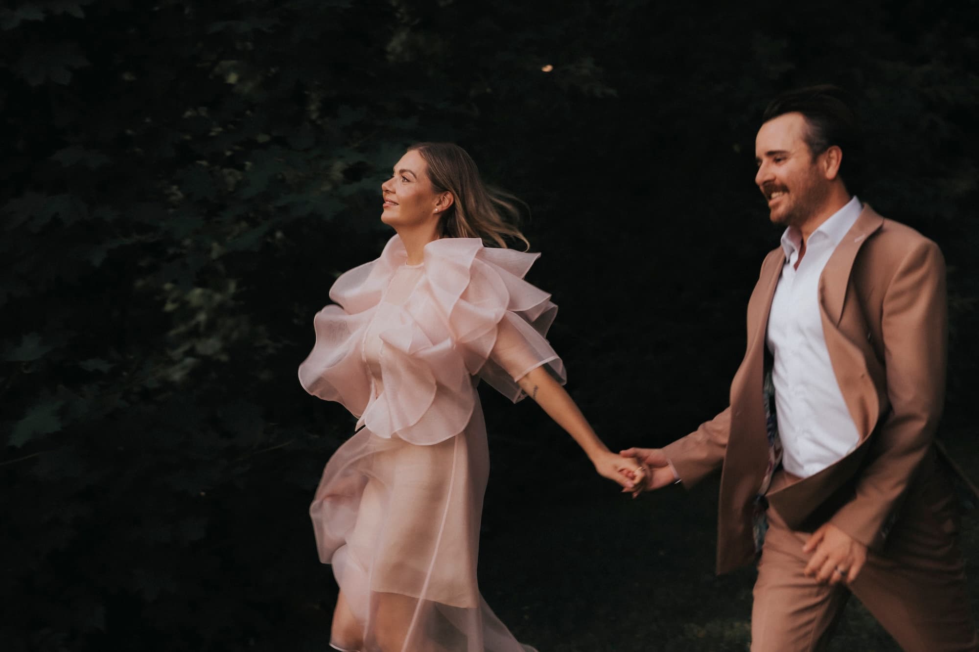 A couple joyfully holding hands while walking outdoors. The woman, in a flowing Flora Dress by Noémiah, features an embroidered neckline, and the man in a brown suit are surrounded by lush green foliage. Both are smiling as they move forward, embracing the beauty of their handmade-to-order attire.