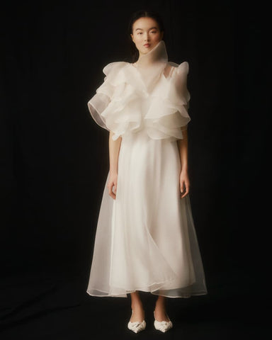 Against a dark background, a woman is seen wearing the Flora Dress by Noémiah. This elegant garment is crafted from white silk organza, featuring ruffled sleeves and a high, embroidered neckline. The dress has a semi-transparent design that exudes a soft, ethereal charm. Her hair is pulled back, and she completes her look with white shoes.