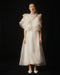 Against a dark background, a woman is seen wearing the Flora Dress by Noémiah. This elegant garment is crafted from white silk organza, featuring ruffled sleeves and a high, embroidered neckline. The dress has a semi-transparent design that exudes a soft, ethereal charm. Her hair is pulled back, and she completes her look with white shoes.