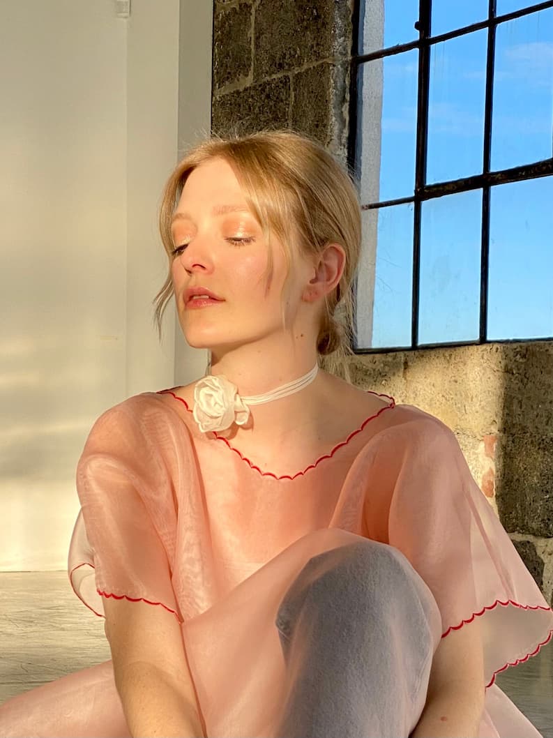 With their eyes closed, someone luxuriates in the sunlight streaming through a window. They're wearing a translucent pink silk organza top with red trim, accompanied by the Noémiah Floral Choker. The room is characterized by its textured wall and expansive window, which casts gentle shadows.