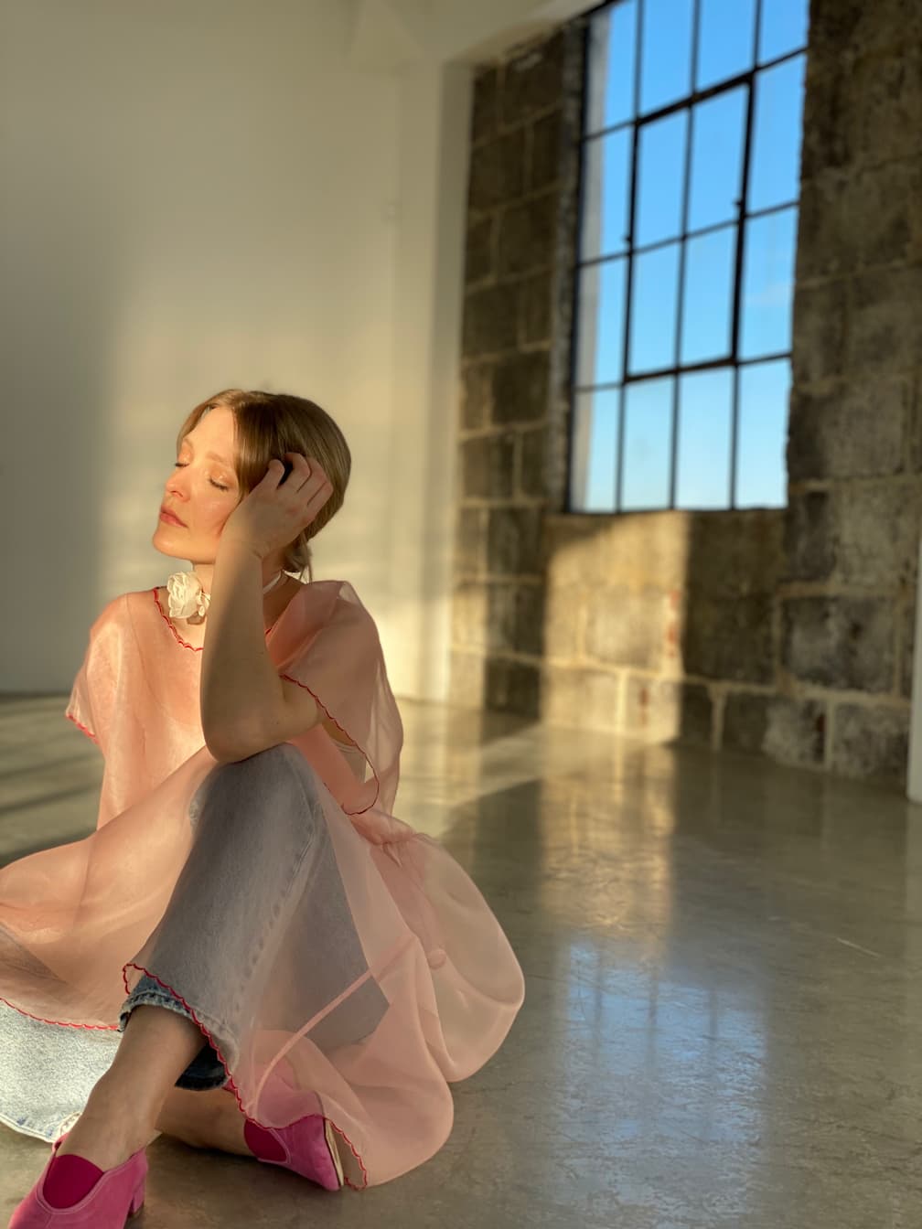 Amidst a peaceful setting, a person sits on a sunlit floor, exuding elegance in a delicately hand-sewn sheer pink dress layered over jeans and complemented by pink shoes. Their light hair gently frames their serene face as they touch it softly with closed eyes. Large windows and an impressive stone wall create the tranquil backdrop for this scene, enhanced by the subtle charm of the Noémiah Floral Choker around their neck.