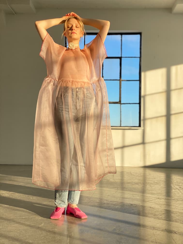 In a sunlit room, a person stands with arms raised and eyes closed, wearing a sheer pink silk organza dress over blue jeans and pink shoes. Sunlight streams through a large window, casting shadows as they model the look complemented by the Floral Choker from Noémiah.