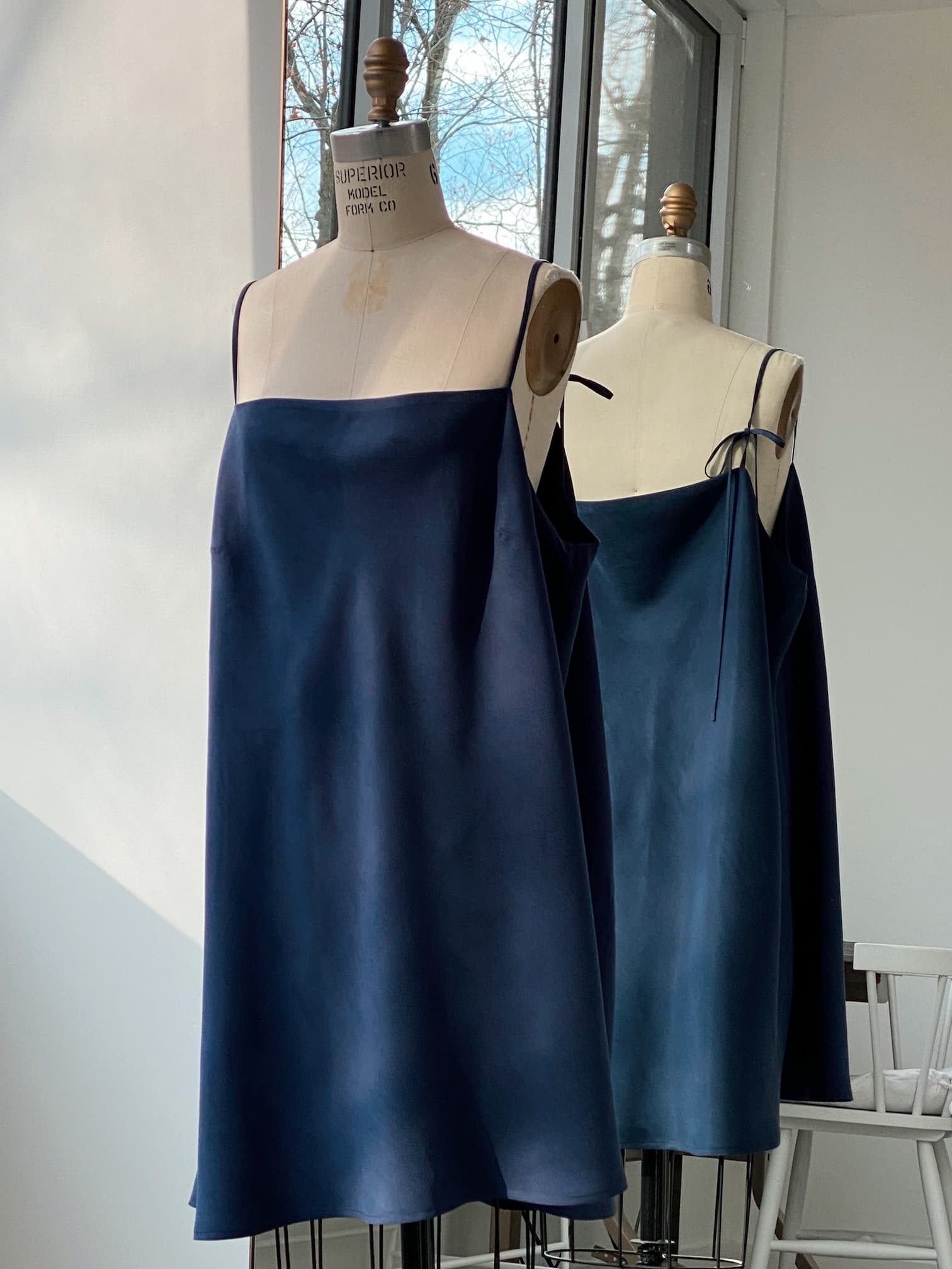 A mannequin showcases the Florence Dress — Lyocell by Noémiah in a navy blue shade, its reflection captured in a nearby mirror. This eco-friendly dress is designed with thin straps and offers a relaxed fit, set against the backdrop of a well-lit room with large windows.