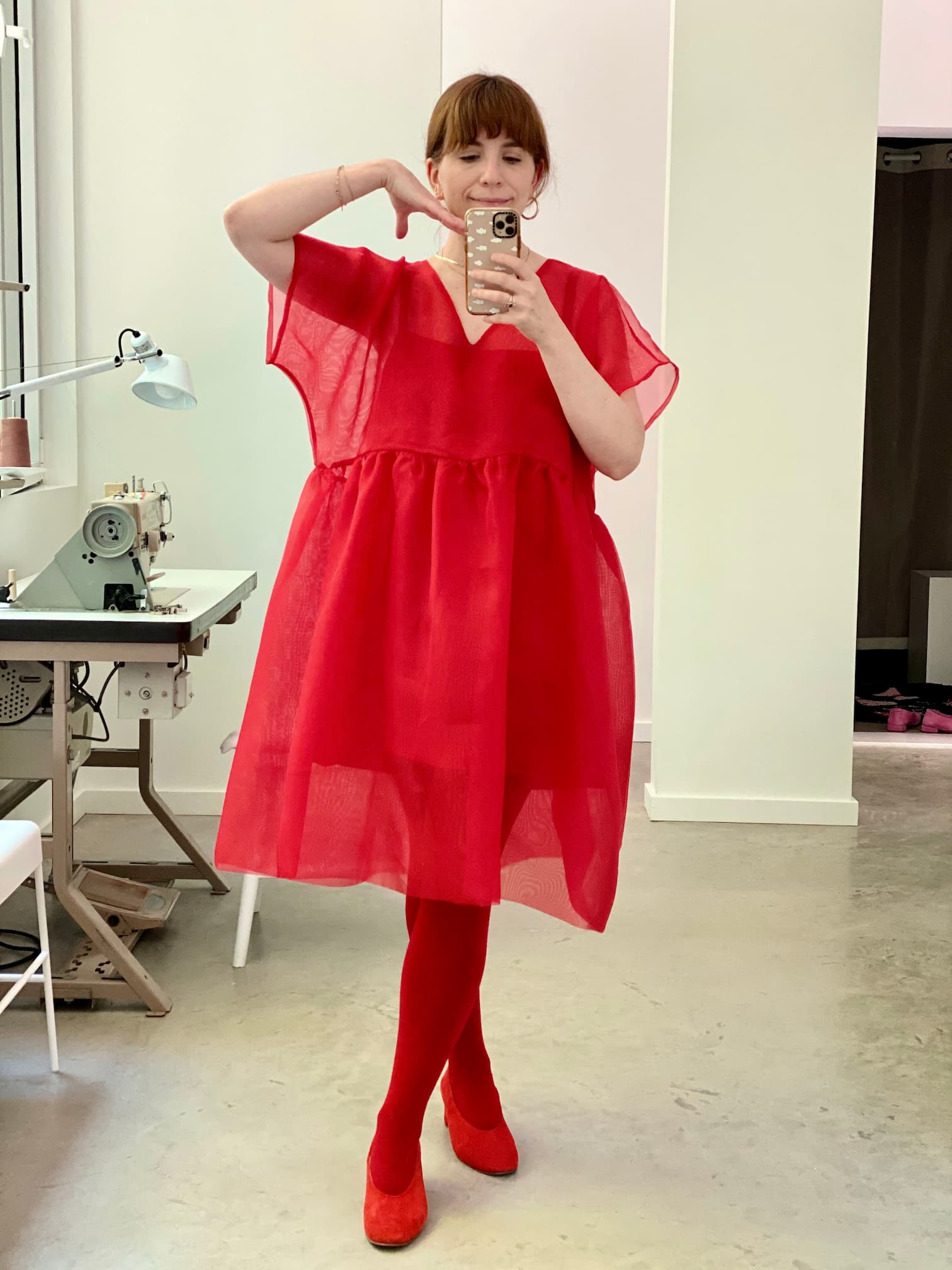 A person stands in front of a mirror taking a selfie, wearing a relaxed fit Florence Dress — Silk by Noémiah, paired with matching red tights and shoes. They are posed on one leg with the other bent in a well-lit room that features a sewing machine in the background.