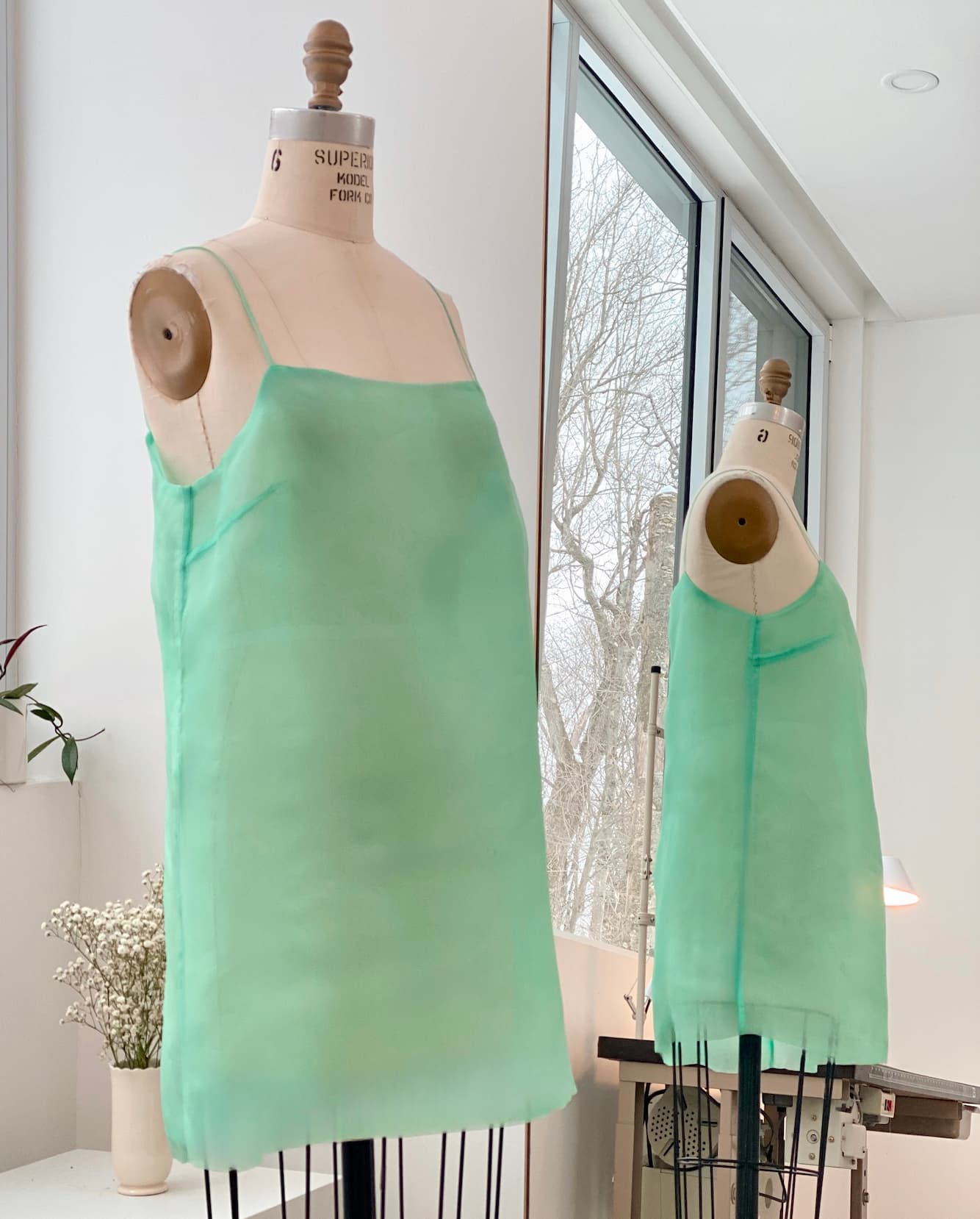 A silk Florence Dress by Noémiah in mint green is elegantly displayed on a mannequin in a bright room with large windows. In the background, another mannequin can be seen. A small table topped with a vase of white flowers sits beside the window, perfectly complementing the relaxed fit of this refined piece.