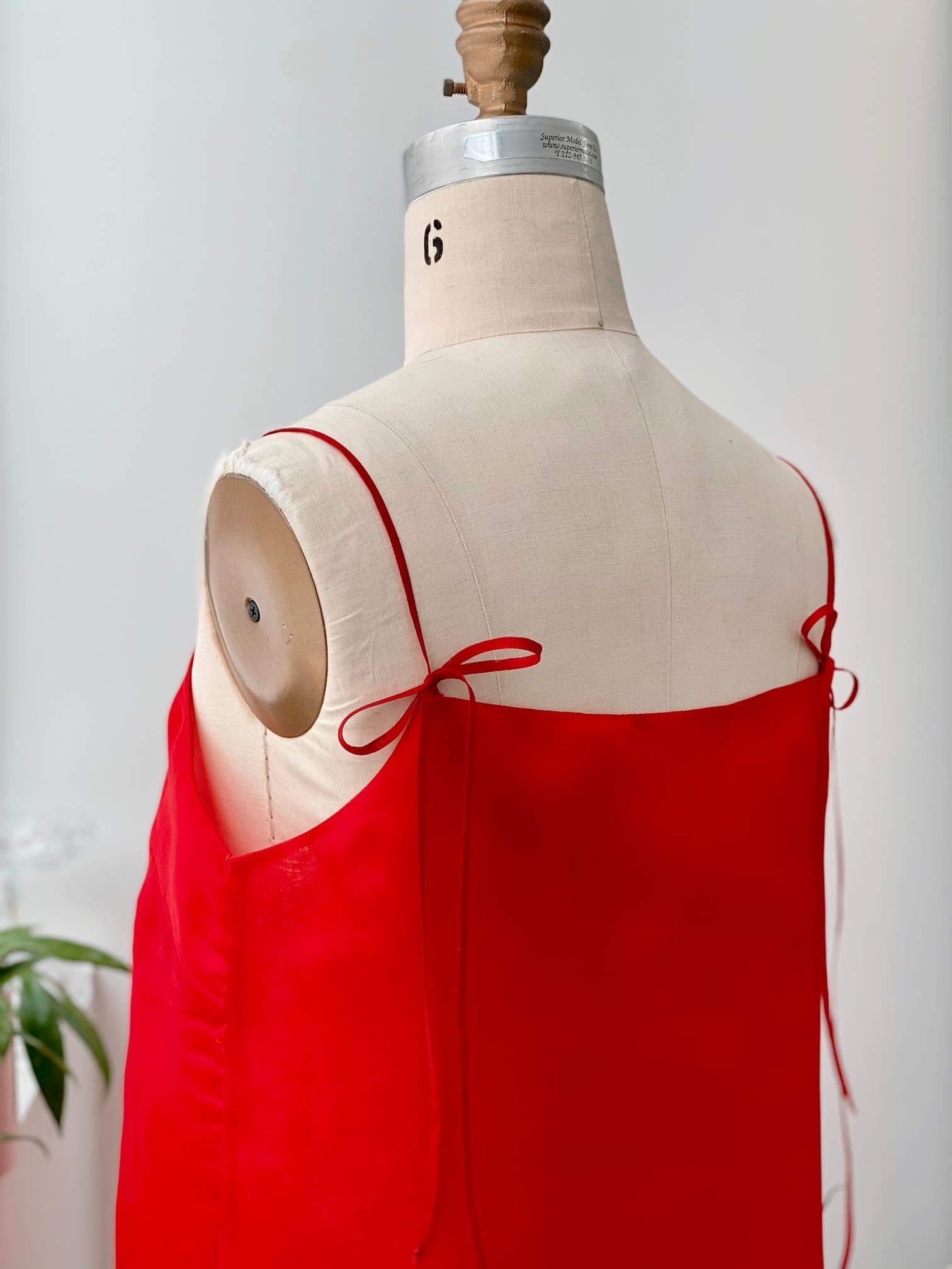 A Florence Dress — Silk by Noémiah in a stunning red hue, featuring delicate straps tied in bows, elegantly adorns a mannequin. The simple yet graceful design of this relaxed fit stands out against a neutral backdrop. A leafy plant subtly enters the scene, infusing nature's charm into the display.