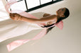 A person stands near a window wearing the Florence Dress by Noémiah, made from sustainable Lyocell. This long, white slip dress features a large pink ribbon tied at the back, creating an elegant atmosphere in the softly lit room.