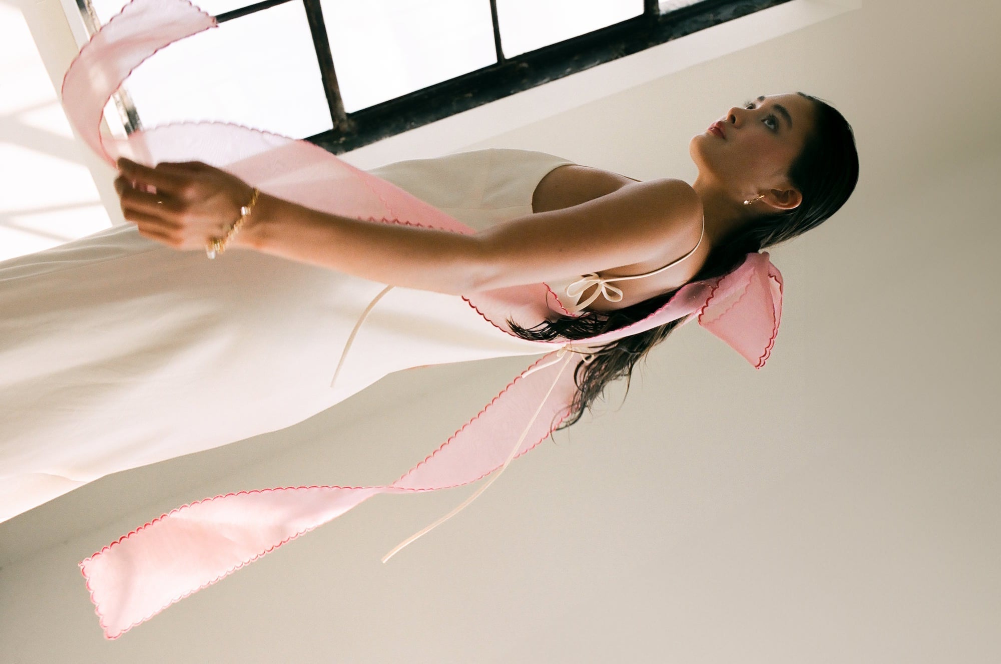 A person stands near a window wearing the Florence Dress by Noémiah, made from sustainable Lyocell. This long, white slip dress features a large pink ribbon tied at the back, creating an elegant atmosphere in the softly lit room.