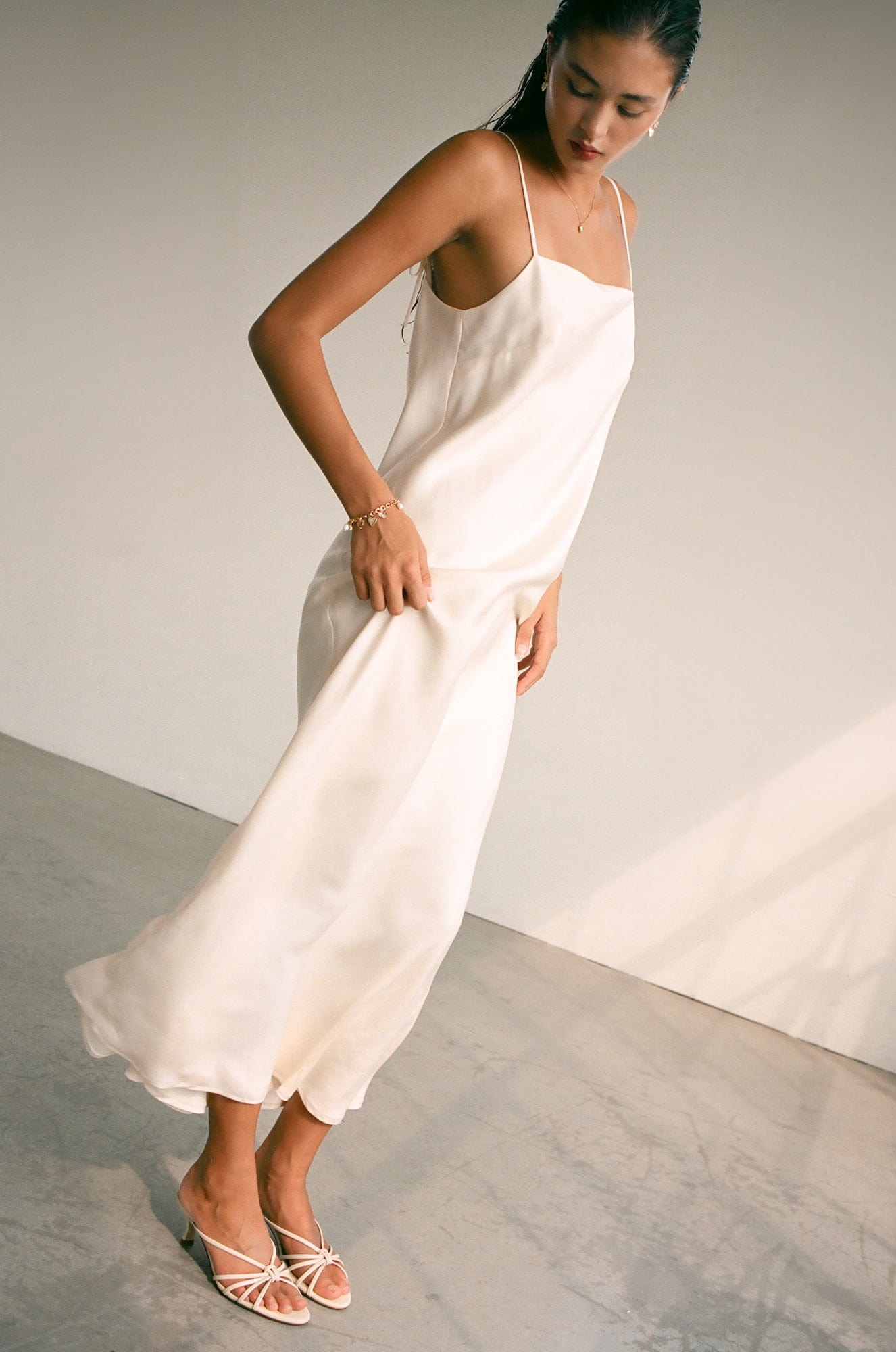 A woman wearing the Florence Lyocell Dress — Bridal Edit by Noémiah poses against a minimalistic backdrop. Crafted from Lyocell, this sustainable slip dress enhances her look with strappy sandals and pulled-back hair. The soft lighting creates an elegant and serene atmosphere as she gazes downwards.