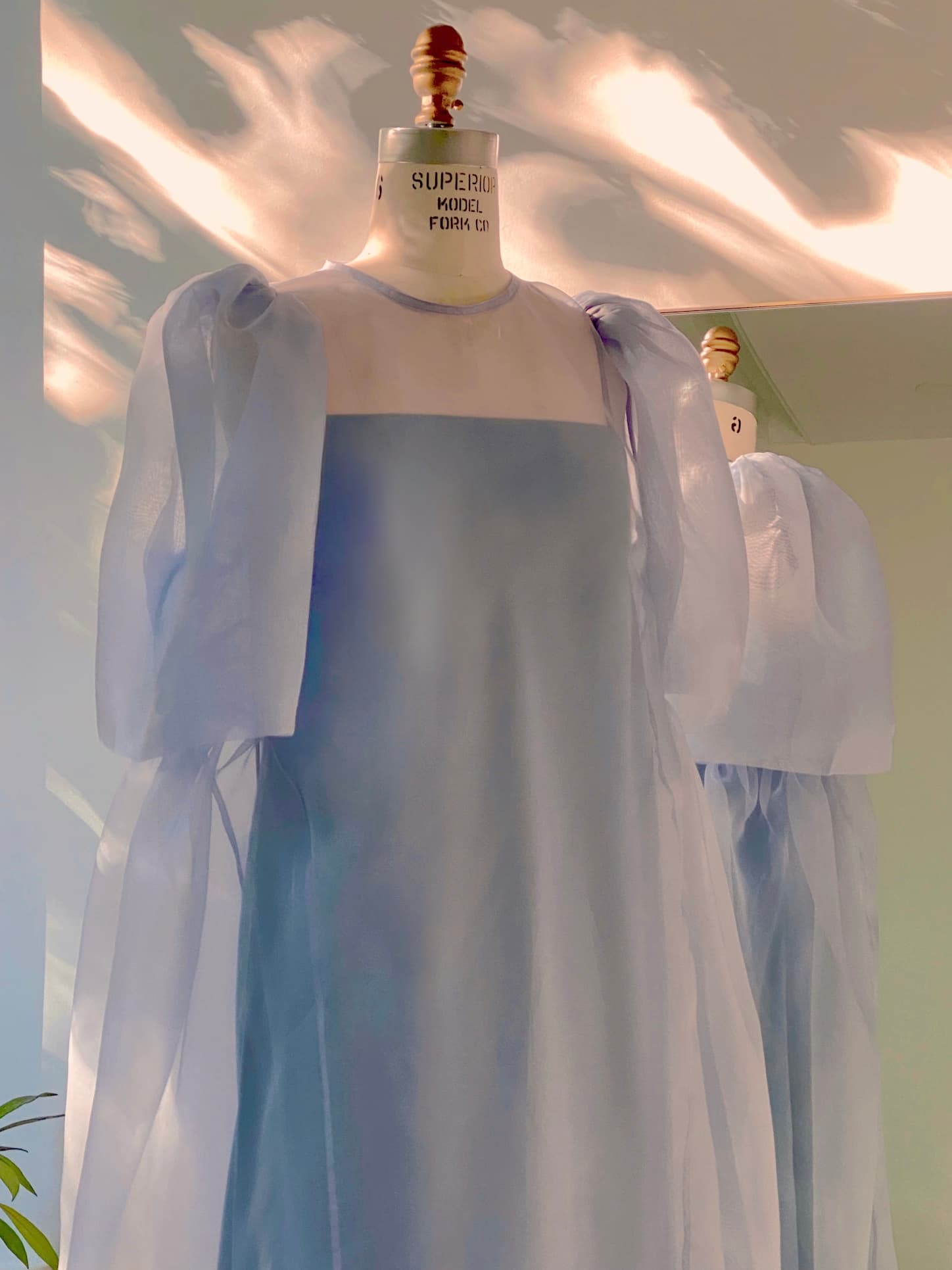 Two mannequins showcase Noémiah's Florence Lyocell Dress — Classic in a minimalist room, their sheer, pale blue fabric with puffed sleeves and square necklines bathed in gentle, dappled sunlight.